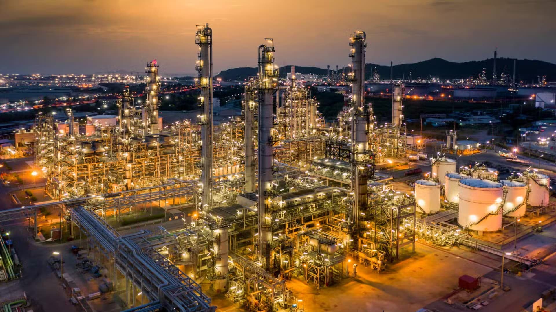 La raffineria al tramonto, vista dall’alto. Le unità di processo illuminate, il parco serbatoi sulla destra e le luci del polo industriale fino all’orizzonte.